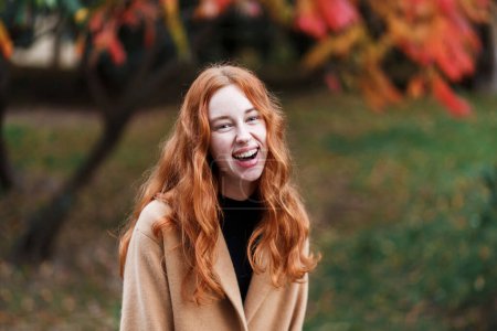 Téléchargez les photos : Heureuse femme aux cheveux roux dans un manteau marche dans un parc ensoleillé en automne - en image libre de droit
