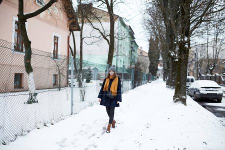 Téléchargez les photos : Femme en écharpe jaune et manteau bleu marche seule dans la rue enneigée - en image libre de droit
