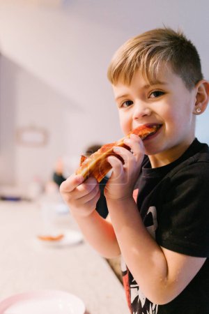 Photo for Smiling white boy taking bite of pizza staring at camera - Royalty Free Image