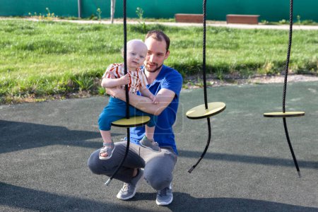 Téléchargez les photos : Papa tient son fils sur une balançoire ronde sur une corde dans la cour de récréation - en image libre de droit