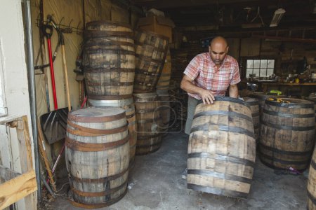 Téléchargez les photos : Un homme roule un grand tonneau de bourbon de bois d'une pile dans un garage - en image libre de droit
