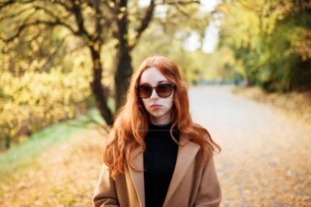 Téléchargez les photos : Élégante femme aux cheveux roux dans un manteau marche dans un parc ensoleillé en automne - en image libre de droit