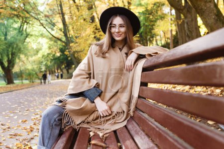 Téléchargez les photos : Femme aux cheveux bruns dans un chapeau noir et poncho sur un banc en automne - en image libre de droit