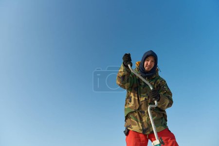 Foto de Un chico asiático calurosamente vestido perfora un agujero en el hielo en la pesca de invierno - Imagen libre de derechos