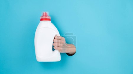 Kobieta trzyma białą butelkę detergentu. Ręka kobiet wystająca z niebieskiego papieru w tle
