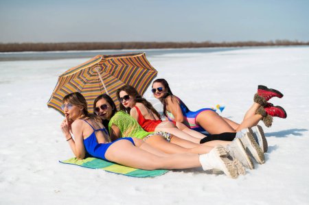 Foto de Cuatro amigas en traje de baño se esconden bajo un paraguas del sol en una playa nevada. Chicas calientes posando en bikinis al aire libre en invierno - Imagen libre de derechos