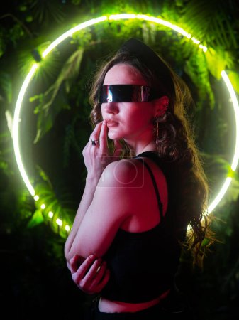 Téléchargez les photos : Femme caucasienne en lunettes de soleil panoramiques sur le fond d'une lampe au néon annulaire dans les plantes - en image libre de droit
