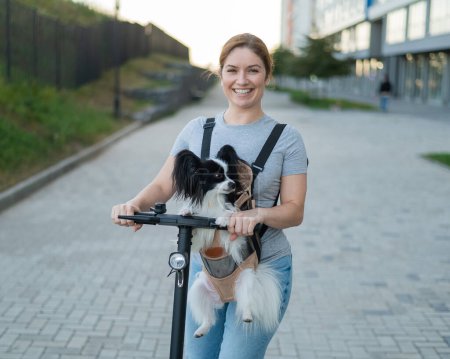 Una mujer monta un scooter eléctrico con un perro en una mochila. Pappilion Spaniel Continental en una honda