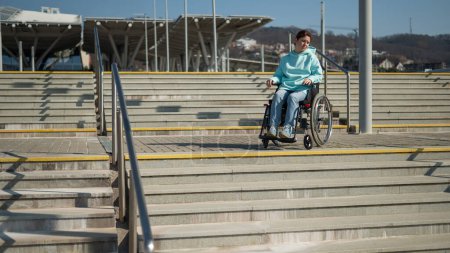 Photo for A caucasian woman in a wheelchair cannot go down stairs without a ramp. Hard to access environment - Royalty Free Image