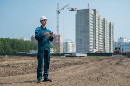 Photo for A man in a helmet and overalls controls a drone at a construction site. The builder carries out technical oversight - Royalty Free Image