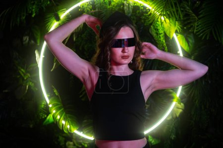 Téléchargez les photos : Femme caucasienne en lunettes de soleil panoramiques sur le fond d'une lampe au néon annulaire dans les plantes - en image libre de droit