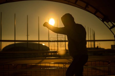 Foto de Un hombre con una sudadera entrena en boxeo en el estadio al atardecer. Silueta de atleta - Imagen libre de derechos