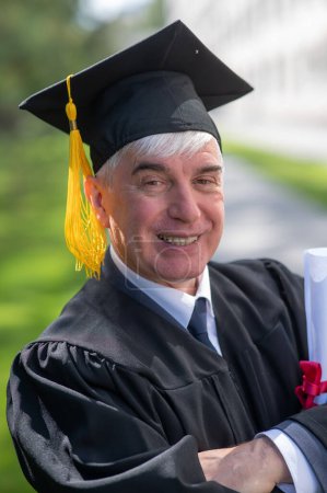 Photo for Portrait of an elderly man in a graduation gown and with a diploma in his hands outdoors. Vertical - Royalty Free Image
