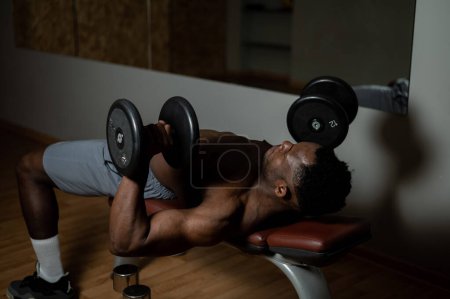 Photo for Shirtless afro american man doing exercises with dumbbells lying on bench in gym - Royalty Free Image
