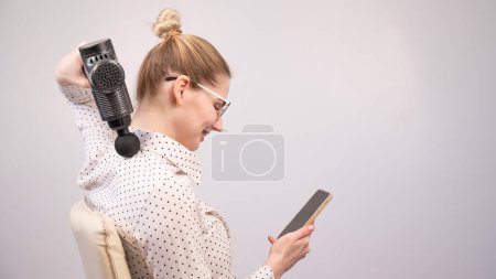 Téléchargez les photos : Une jeune femme s'assoit sur une chaise, utilise un smartphone et se fait masser avec un pistolet électro massage - en image libre de droit