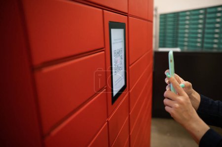 Photo for A woman scans a red code to pick up a parcel at a parcel machine. Automated Postal Box - Royalty Free Image