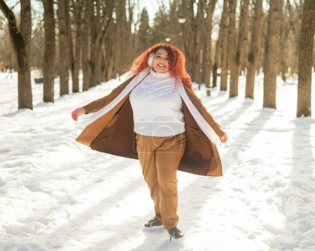 Téléchargez les photos : Une grosse femme écoute de la musique avec des écouteurs et danse dans le parc en hiver - en image libre de droit