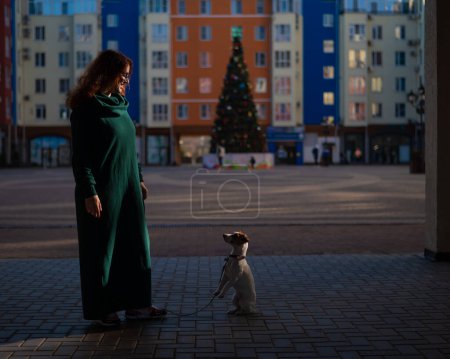 Red-haired woman in a green dress with a dog Jack Russell Terrier in the square against the background of the Christmas tree