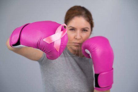 Téléchargez les photos : Femme blanche en gants de boxe roses avec un ruban rose sur la poitrine sur un fond gris. Lutte contre le cancer du sein - en image libre de droit