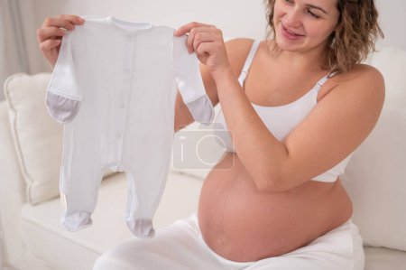 Téléchargez les photos : Une femme enceinte s'assoit sur un canapé blanc et tient des vêtements pour le bébé attendu - en image libre de droit