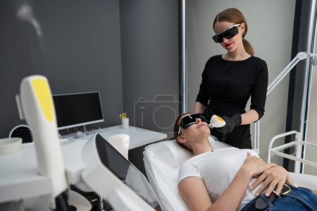 Mujer caucásica joven con gafas en el procedimiento de fotorejuvenecimiento. Médico cosmetólogo con una paciente femenina