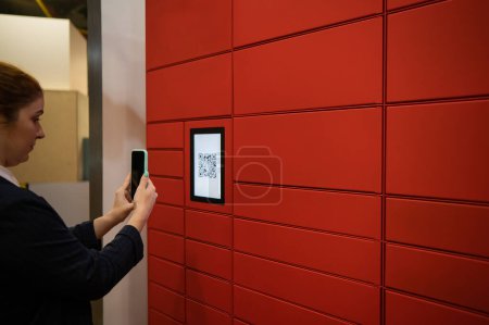 Photo for A woman scans a red code to pick up a parcel at a parcel machine. Automated Postal Box - Royalty Free Image
