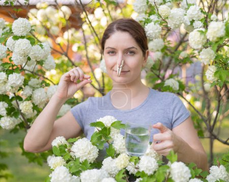 Foto de Mujer caucásica toma un medicamento antihistamínico y quita una pinza de su nariz cerca de un árbol con flores - Imagen libre de derechos