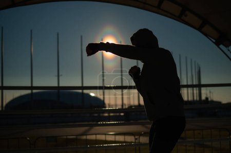 Foto de Un hombre con una sudadera entrena en boxeo en el estadio al atardecer. Silueta de atleta - Imagen libre de derechos