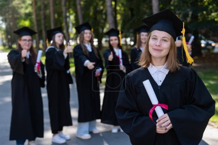 Photo for Group of happy students in graduation gowns outdoors. A young girl with a diploma in her hands in the foreground - Royalty Free Image