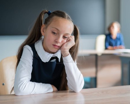 Little caucasian girl is bored at the lesson at school. The schoolgirl is sitting at her desk and the teacher is sitting in the background