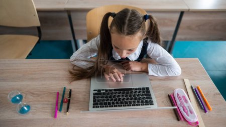 Foto de Chica caucásica se sienta en un escritorio en la escuela y estudia en un ordenador portátil. Reloj de arena sobre la mesa - Imagen libre de derechos