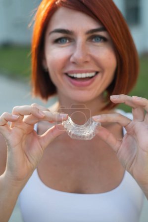 Photo for Red-haired Caucasian woman holding transparent mouthguards for bite correction outdoors. A girl with a beautiful snow-white smile uses silicone braces. - Royalty Free Image