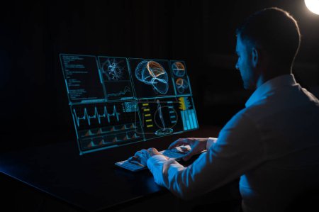 Téléchargez les photos : Un homme caucasien est assis dans le noir devant un menu virtuel et tape sur un clavier. recherche sur le cerveau humain - en image libre de droit