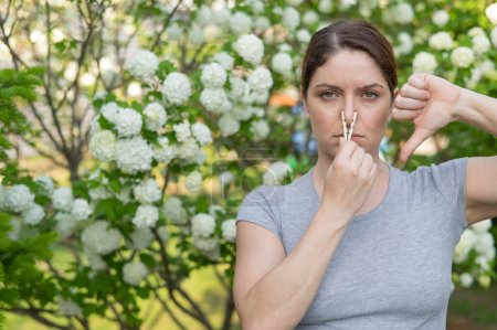 Téléchargez les photos : Une femme souffre d'une pince à linge sur son nez et montre un pouce vers le bas sur une promenade dans un parc fleuri. Allergie - en image libre de droit