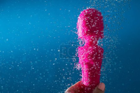 A woman washes a pink vibrator under running water on a blue background. Sex toy hygiene concept