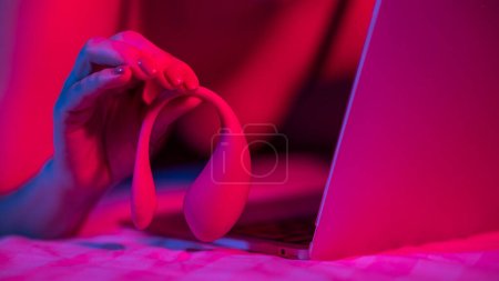 Téléchargez les photos : Une femme est allongée dans son lit tenant un vibromasseur incurvé et regardant un ordinateur portable. Fille en utilisant un jouet sexuel dans la lumière bleu-rouge - en image libre de droit