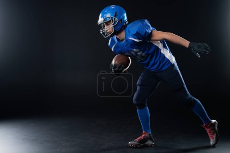 Photo for Full length portrait of a man in a blue american football uniform against a black background. Sportsman in a helmet with a ball - Royalty Free Image