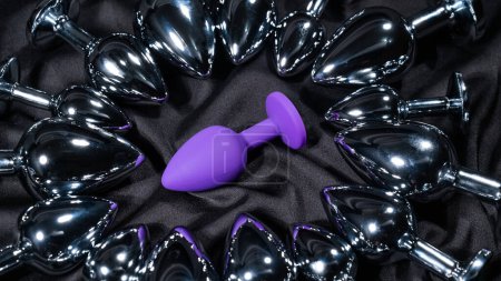 Foto de Purple silicone butt plug among many metal ones on a black silk sheet - Imagen libre de derechos
