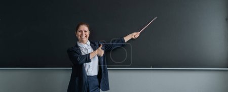 Die rothaarige Kaukasierin im Strumpfanzug zeigt den Daumen nach oben. Lächelnde Lehrerin mit einem Zeiger auf die Tafel. Breitbild
