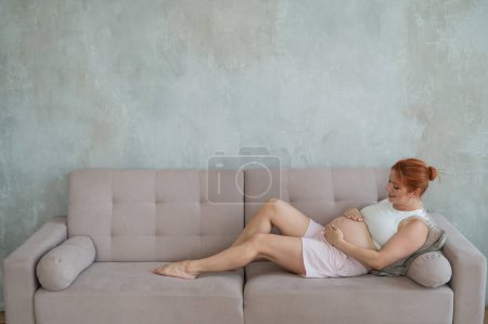 Photo for Caucasian pregnant woman lies on the sofa, strokes and communicates with her tummy - Royalty Free Image