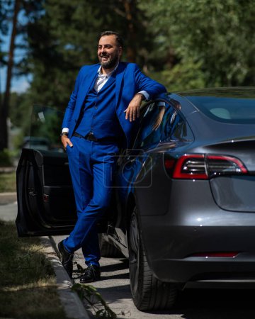 Photo for Caucasian bearded man in a blue suit gets out of a black electro car in the countryside in summer - Royalty Free Image