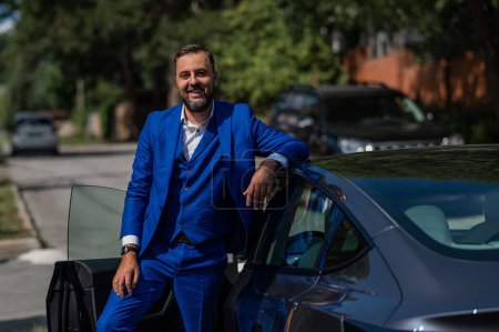 Photo for Caucasian bearded man in a blue suit gets out of a black electro car in the countryside in summer - Royalty Free Image
