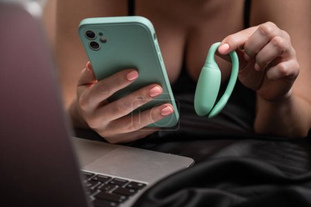 A woman lies on a bed and synchronizes the kegel machine with a smartphone. Girl watching video on laptop