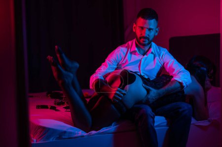 Bearded caucasian man spanks a woman with a leather whip while sitting on a bed in neon light. BDSM sex preferences