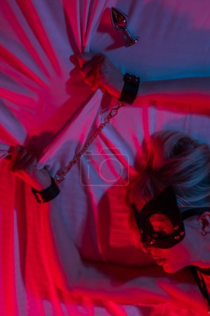 Foto de Mujer rubia sexy con una máscara de cuero y esposas y un collar se encuentra en el dormitorio en una luz de neón rojo-azul. Juegos de rol - Imagen libre de derechos
