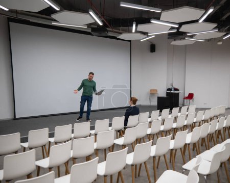 A red-haired Caucasian businesswoman sits in the front row of an empty conference room. Bearded man giving a lecture