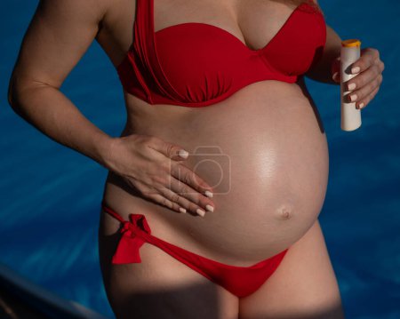 Photo for Close-up of the belly of a pregnant woman sunbathing in a red bikini. Expectant mother applies sunscreen - Royalty Free Image