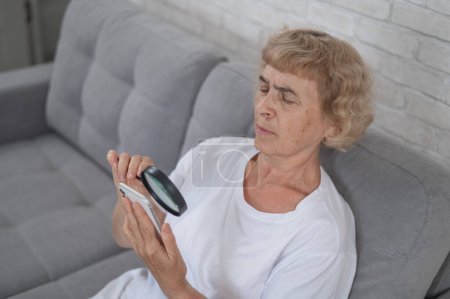 Foto de Anciana mujer caucásica con miopía y tratando de leer un mensaje en un teléfono inteligente con una lupa - Imagen libre de derechos