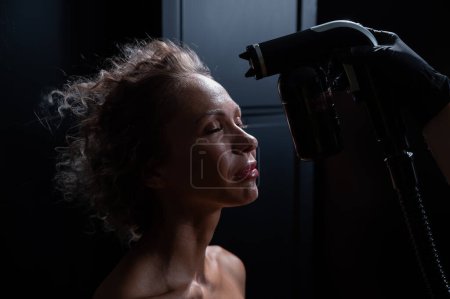 Foto de Un maestro aplica pintura tintada a la cara de una mujer caucásica. Procedimiento de bronceado instantáneo. - Imagen libre de derechos