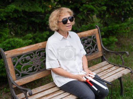 An elderly blind woman sits on a bench in the park with a folded tactile cane in her hands